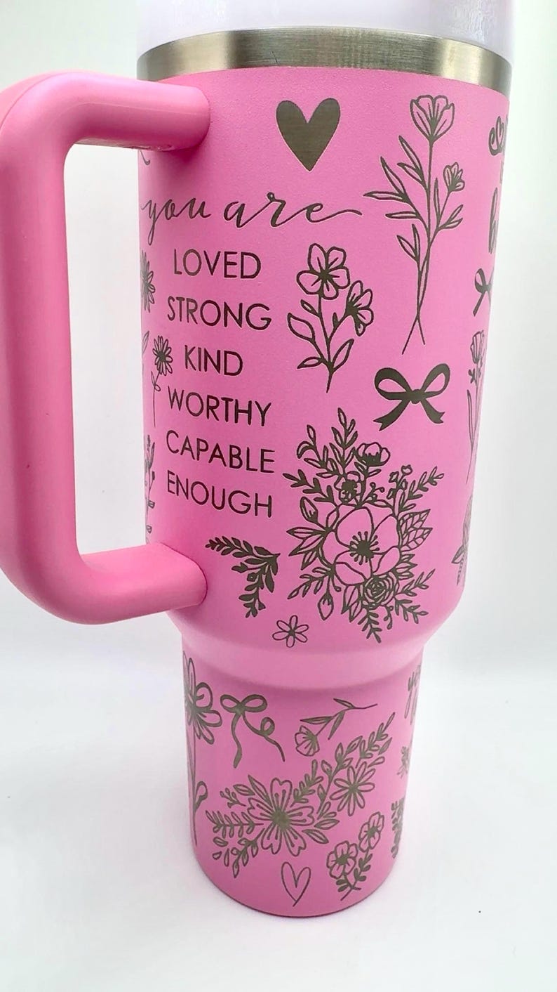 Peut inclure: Gobelet isotherme rose avec une poign&eacute;e, orn&eacute; de motifs floraux et de n&oelig;uds. Le gobelet porte les mots "you are LOVED STRONG KIND WORTHY CAPABLE ENOUGH" en &eacute;criture cursive. Comprend un symbole de c&oelig;ur.
