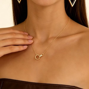 Puede incluir: Un collar dorado con un colgante circular y un cierre. El colgante tiene grabado el nombre "Olivia". El collar se lleva alrededor del cuello. En el fondo se ven pendientes triangulares dorados.
