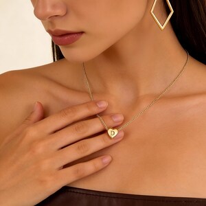 Puede incluir: Un collar dorado con un colgante en forma de corazón con la letra "D". El collar lo lleva una persona, y el colgante está centrado. La persona también lleva pendientes dorados en forma de diamante.