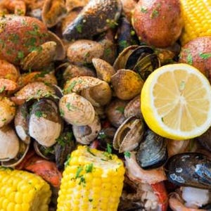 May include: A close-up shot of a seafood boil featuring mussels, clams, shrimp, potatoes, and corn on the cob. A lemon wedge adds a bright yellow accent to the dish. The ingredients are seasoned with herbs and spices.