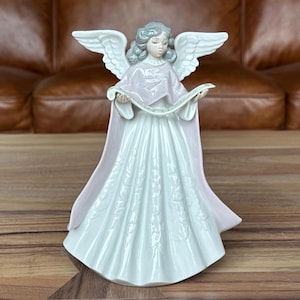 May include: A ceramic angel figurine with outstretched wings and a flowing gown. The angel has grey hair and is holding a scroll. The gown is white with a floral pattern, and the cape is pale pink. The figurine is on a wooden surface.