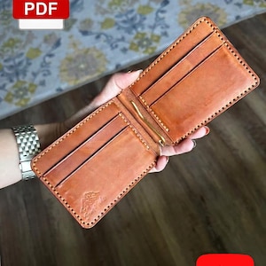 May include: A tan leather bifold wallet featuring a central gold-toned metal money clip, multiple interior card slots, and a small embossed mountain logo on the bottom left corner. The wallet is constructed with visible tan stitching along the edges.