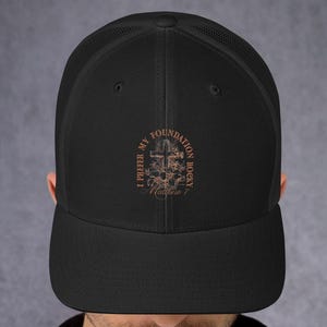 May include: Black trucker hat with a curved brim and mesh back. The hat features a brown graphic with the text "I PREFER MY FOUNDATION ROCKY Matthew 7". The hat is worn by a person.