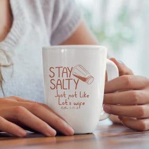 Może przedstawiać: Biały ceramiczny kubek z napisem "STAY SALTY" i grafiką solniczki. Kubek zawiera również tekst "Just not like Lot's wife" i odniesienie do wersetu biblijnego. Kubek jest trzymany przez osobę.