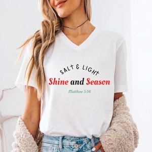 May include: White V-neck t-shirt with the text "SALT & LIGHT Shine and Season Matthew 5:14" in black and red. The shirt is a casual style, suitable for everyday wear.