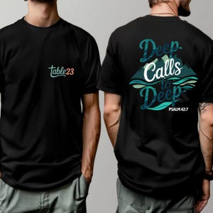 Puede incluir: Camiseta negra con el texto "table23" en verde azulado y rosa en la parte delantera. La parte trasera de la camiseta presenta el texto "Deep Calls to Deep" con un diseño de montaña y agua en verde azulado y blanco. Salmo 42:7 está impreso debajo.
