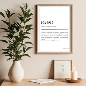 May include: A framed print with the word "POMAPOO" and its pronunciation, along with a definition. The print is in a light wood frame, and there is a vase with greenery to the left. A smaller framed print and a candle are also visible.