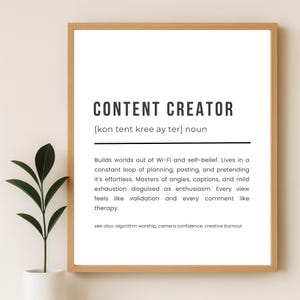 Puede incluir: Impresión enmarcada con las palabras "CONTENT CREATOR" en negrita negra. La definición describe el rol, incluyendo planificación, publicación y fingir que es sin esfuerzo. Marco marrón claro, planta en maceta blanca a la izquierda.