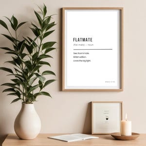 May include: A framed print with the word "FLATMATE" in black text. Below the word is a definition, "See: Roommate. British edition. Loves the big light." A vase with greenery is to the left. A smaller framed print and a candle are below.