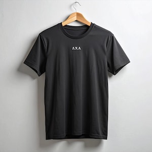 May include: A black short-sleeve t-shirt with the letters "AXA" printed in white on the chest. The shirt is hanging on a wooden hanger against a white background. The t-shirt has a crew neck.