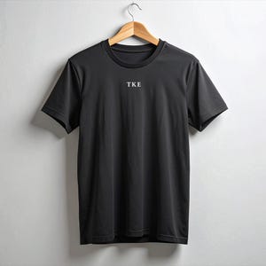 May include: A black crew neck t-shirt hanging on a wooden hanger. The shirt has short sleeves and the letters "TKE" are printed in white on the chest.