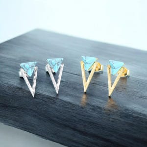 Könnte beinhalten: Zwei Paar Ohrstecker, eines in Silber und eines in Gold. Jeder Ohrring hat einen türkisfarbenen Dreiecksstein, der in einem V-förmigen Rahmen gefasst ist. Die Ohrringe werden auf einer dunkelgrauen Oberfläche präsentiert.