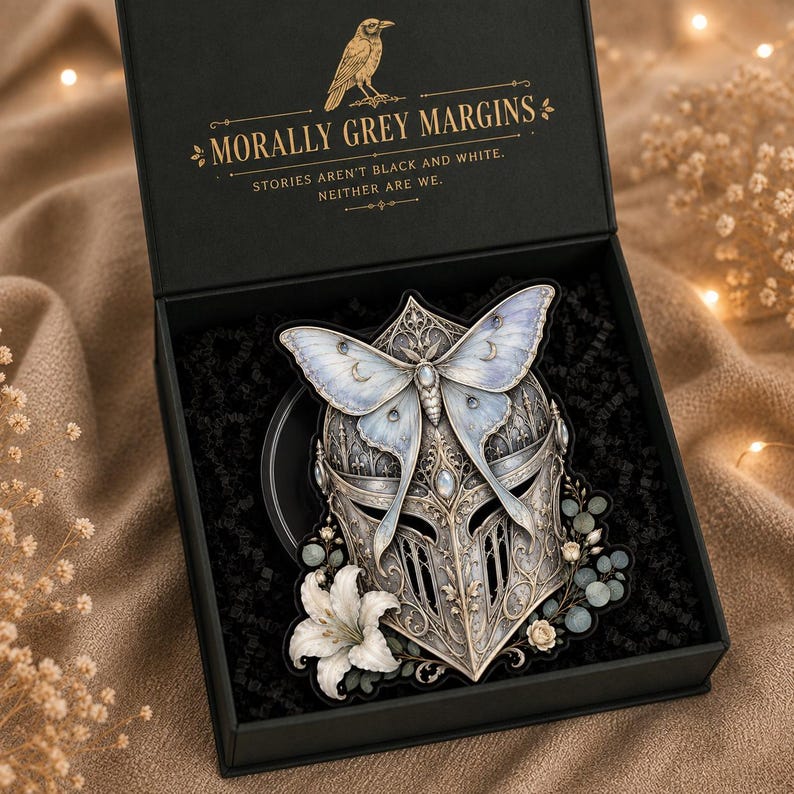 May include: A black gift box containing a decorative knight helmet ornament adorned with a large pale blue moth, white lily flowers, and small green leaves. The box lid features a gold raven icon and the text MORALLY GREY MARGINS.