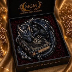 May include: A black box containing a dark blue dragon figurine resting on a book with gold accents. The box lid features the text MGM MORALLY GREY MARGINS, and the base is printed with MADE BY HAND. MADE WITH HEART. The dragon has gold horns.