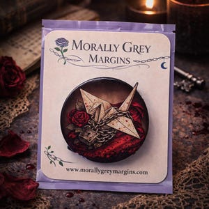 May include: A round pin featuring a paper crane, roses, and a chain with a padlock. The pin is packaged in a clear plastic sleeve with the text "Morally Grey Margins" and the website address www.morallygreymargins.com.