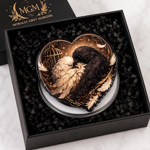 May include: A heart-shaped decorative item featuring an illustration of a white bird and a black bird nestled together near a golden birdcage. The piece is presented in a black gift box filled with black paper filler, with the text MGM MORALLY GREY MARGINS printed on the box lid.