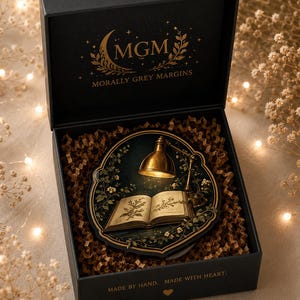 May include: A black gift box containing a circular decorative plaque featuring an open book illuminated by a brass desk lamp, surrounded by botanical illustrations. The box lid displays the gold text MGM MORALLY GREY MARGINS and MADE BY HAND. MADE WITH HEART.