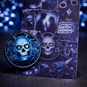 Puede incluir: Un conjunto de pines y tarjetas azules y negras con diseños de calaveras. El pin tiene una calavera con ojos tachados y ojos en forma de corazón, rodeada de rosas y espadas. La tarjeta tiene el texto "COUPLES WHO SLAY TOGETHER STAY TOGETHER".