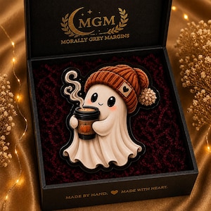 May include: A black gift box containing a sticker of a white ghost wearing a rust-colored knit beanie and holding a black coffee cup with a pumpkin graphic. The box lid features the gold text MGM MORALLY GREY MARGINS and MADE BY HAND.