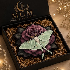 May include: A black gift box containing a decorative item featuring a large, dark purple rose with dark green leaves and a pale mint green moth with gold crescent moon accents. The box lid displays the gold text MGM MORALLY GREY MARGINS.