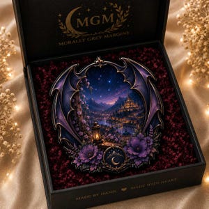 May include: A circular decorative plaque featuring a fantasy landscape with a castle, mountains, and a night sky, framed by purple bat-like wings and flowers. It is presented in a black box with the text MGM MORALLY GREY MARGINS inside.