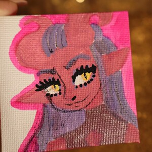 May include: A small, square canvas painting featuring a cartoon character with pink skin, purple hair, and yellow eyes. The character has large eyelashes and a small smile. The background is a bright pink color.