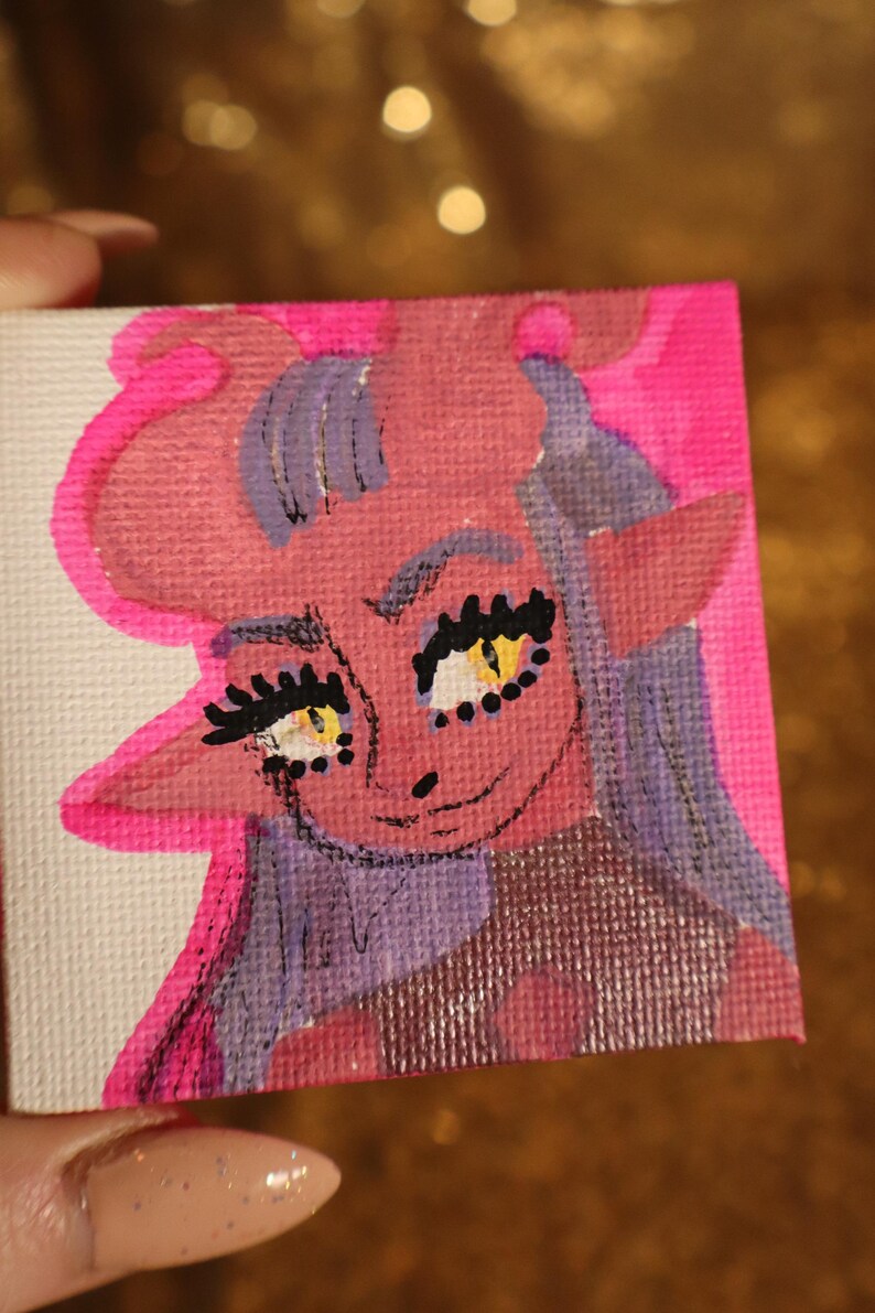 May include: A small, square canvas painting featuring a cartoon character with pink skin, purple hair, and yellow eyes. The character has large eyelashes and a small smile. The background is a bright pink color.