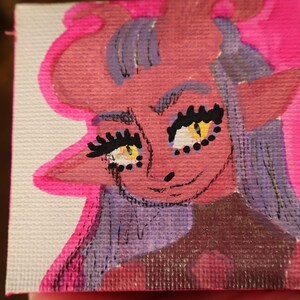 May include: A small, square painting featuring a cartoonish demon with pink skin, purple hair, and yellow eyes. The artwork is outlined in bright pink and held in a hand, with a blurred background.