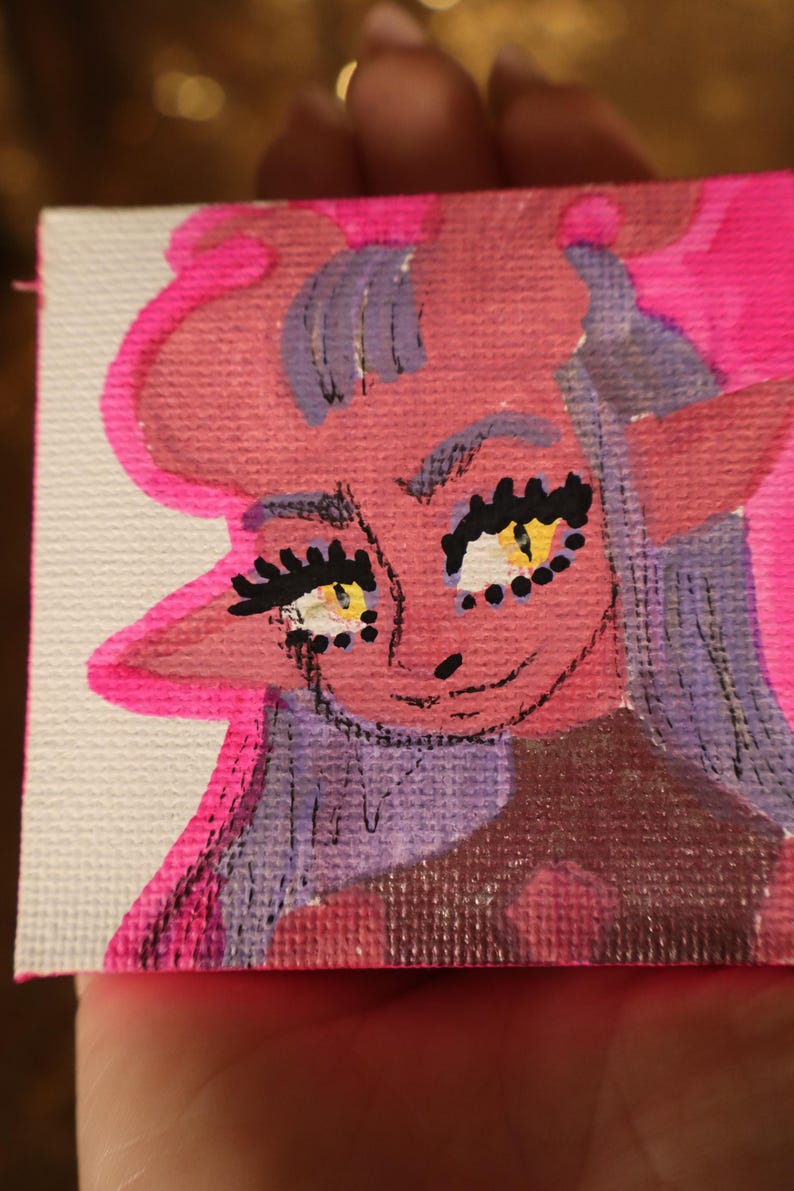 May include: A small, square painting featuring a cartoonish demon with pink skin, purple hair, and yellow eyes. The artwork is outlined in bright pink and held in a hand, with a blurred background.