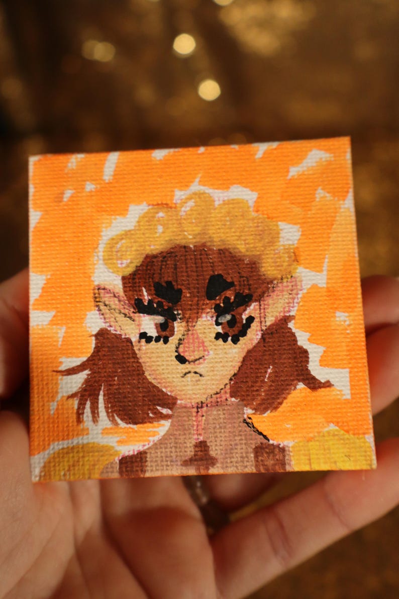 May include: A small square painting featuring a stylized portrait of a person with brown hair and large eyes. The artwork is set against an orange and yellow background. The painting is held in a hand.