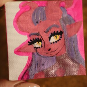 May include: A small, square canvas art piece featuring a cartoonish demon character. The artwork uses pink, red, yellow, and purple hues. The demon has horns, pointed ears, and a smiling expression. The background is a bright pink.