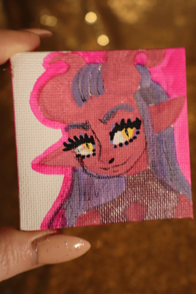 May include: A small, square canvas art piece featuring a cartoonish demon character. The artwork uses pink, red, yellow, and purple hues. The demon has horns, pointed ears, and a smiling expression. The background is a bright pink.