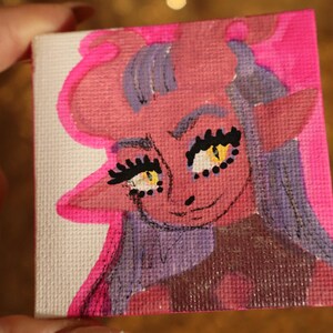 May include: A small, square canvas painting featuring a cartoon character with pink skin, purple hair, and yellow eyes. The character has large eyelashes and a smiling expression. The artwork is outlined in bright pink.