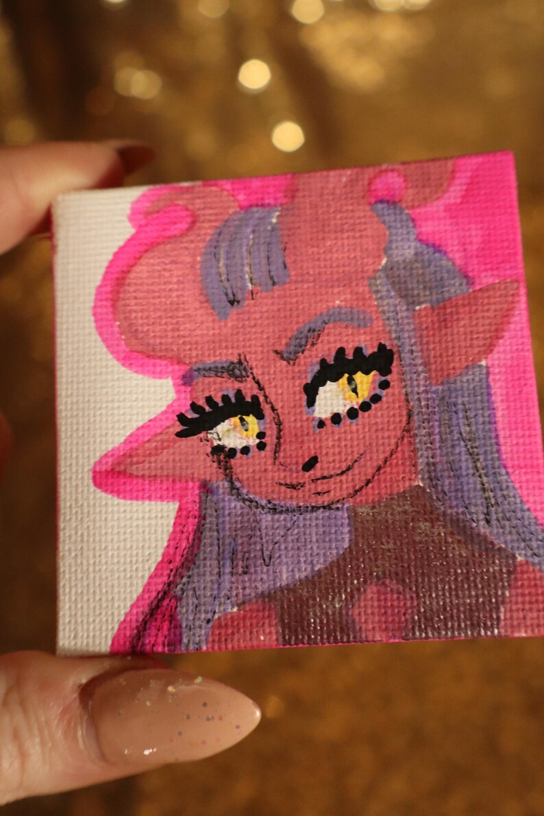 May include: A small, square canvas painting featuring a cartoon character with pink skin, purple hair, and yellow eyes. The character has large eyelashes and a smiling expression. The artwork is outlined in bright pink.