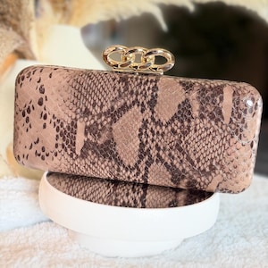 May include: A rectangular clutch bag featuring a brown and black snakeskin pattern. The bag has a smooth, structured exterior and is topped with a gold-toned metal chain link clasp mechanism that serves as the primary closure for the accessory.