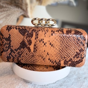 May include: A rectangular clutch bag featuring a brown and tan snakeskin pattern. The bag has a smooth, structured exterior and is topped with a polished gold-tone metal chain link accent that serves as a decorative closure mechanism.