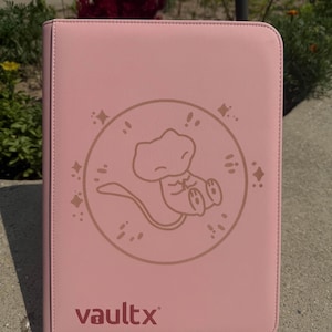 May include: A pink Vaultx trading card binder with a brown illustration of a cartoon character inside a circle. The binder has a zipper closure and the Vaultx logo in the bottom left corner. The background is a concrete surface with greenery.