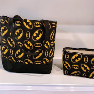 Könnte beinhalten: Eine schwarze Tragetasche und eine passende kleinere Reißverschlusstasche, beide mit einem sich wiederholenden gelb-schwarzen Batman-Logo-Muster. Die Tasche hat schwarze Stoffgriffe und einen einfarbig schwarzen Boden, die kleine Tasche einen Reißverschluss.