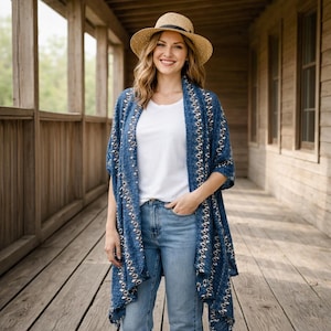 Puede incluir: Una mujer con sombrero de paja, camiseta blanca, vaqueros azules y un chal azul estampado. El chal tiene un diseño color crema en los bordes. La mujer está de pie en un porche de madera.