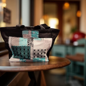 May include: Black tote bag with a patchwork design. The patchwork features squares of teal, gray, and white fabrics with various patterns. The bag has black handles and sits on a wooden table.