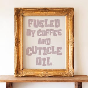 Könnte beinhalten: Ein gerahmter Druck mit dem Text "Fueled by Coffee and Cuticle Oil" in hellem Lila. Der goldfarbene Rahmen ist verziert und steht auf einem Holzregal. Der Hintergrund ist eine schlichte weiße Wand.