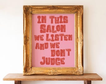 May include: A framed print with the text "IN THIS SALON WE LISTEN AND WE DON'T JUDGE" in red letters on a pink background. The frame is ornate and gold-colored, and the print is displayed on a wooden shelf.