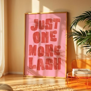 May include: A rectangular wall art print in a gold frame featuring a pink background with the bold red text "JUST ONE MORE LASH" in a rounded, hand-lettered style font. The artwork is displayed in a vertical orientation with a clean, simple design.