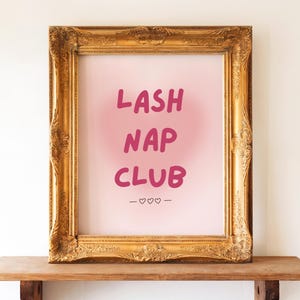 Puede incluir: Una impresión enmarcada con las palabras "LASH NAP CLUB" en letras rosas. La impresión está en un marco dorado adornado y descansa sobre una estantería de madera. Debajo del texto hay tres pequeños símbolos de corazón.