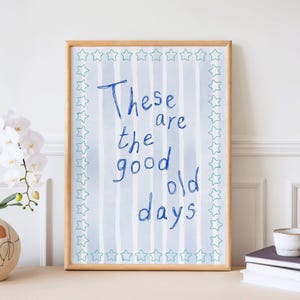 Puede incluir: Un estampado enmarcado con las palabras "These are the good old days" en escritura azul. El fondo tiene rayas azules verticales y un borde de estrellas verdes. El marco es de madera clara.