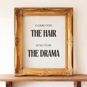 Puede incluir: Una impresión de arte mural rectangular en un marco dorado ornamentado. La impresión blanca presenta texto negro que dice: COME FOR THE HAIR, STAY FOR THE DRAMA. La tipografía utiliza una mezcla de fuentes sans-serif y serif.