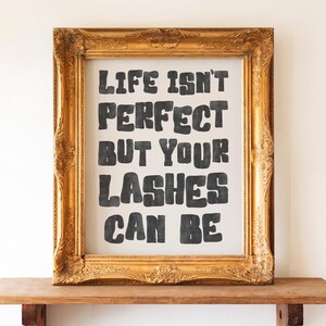 May include: A framed print with the text "LIFE ISN'T PERFECT BUT YOUR LASHES CAN BE" in black letters. The frame is ornate, gold-colored, and sits on a wooden shelf. The background is a neutral color.