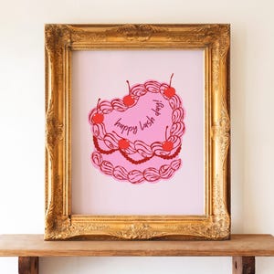 Puede incluir: Una impresión enmarcada con un diseño de pastel rosa en forma de corazón con cerezas rojas y el texto "happy lash day!". La impresión está en un marco dorado adornado, sobre una estantería de madera.