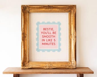 May include: A framed print with the text "BESTIE, YOU'LL BE SMOOTH IN LIKE 5 MINUTES" in red letters on a pink background, surrounded by a blue border. The print is in a gold ornate frame, resting on a wooden surface.