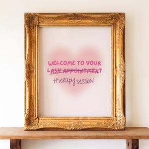 Può includere: Un cartello incorniciato con dettagli dorati ornati. Il cartello recita "BENVENUTO AL TUO APPUNTAMENTO PER LE CIGLIA" con "sessione di terapia" scritto sotto. Lo sfondo ha una forma di cuore rosa.